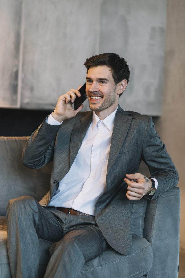 smiling-young-businessman-sitting-sofa-talking-through-mobile-phone smiling-young-businessman-sitting-sofa-talking-through-mobile-phone